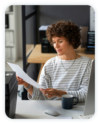 Woman sitting in front of a desktop computer, looking at a piece of paper she's holding in her hands.